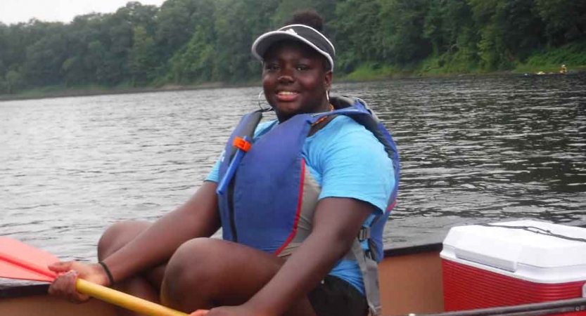 A person wearing a life jacket holds a paddle and smiles for the photo. They are in a boat. There are green trees lining the shore in the background. 